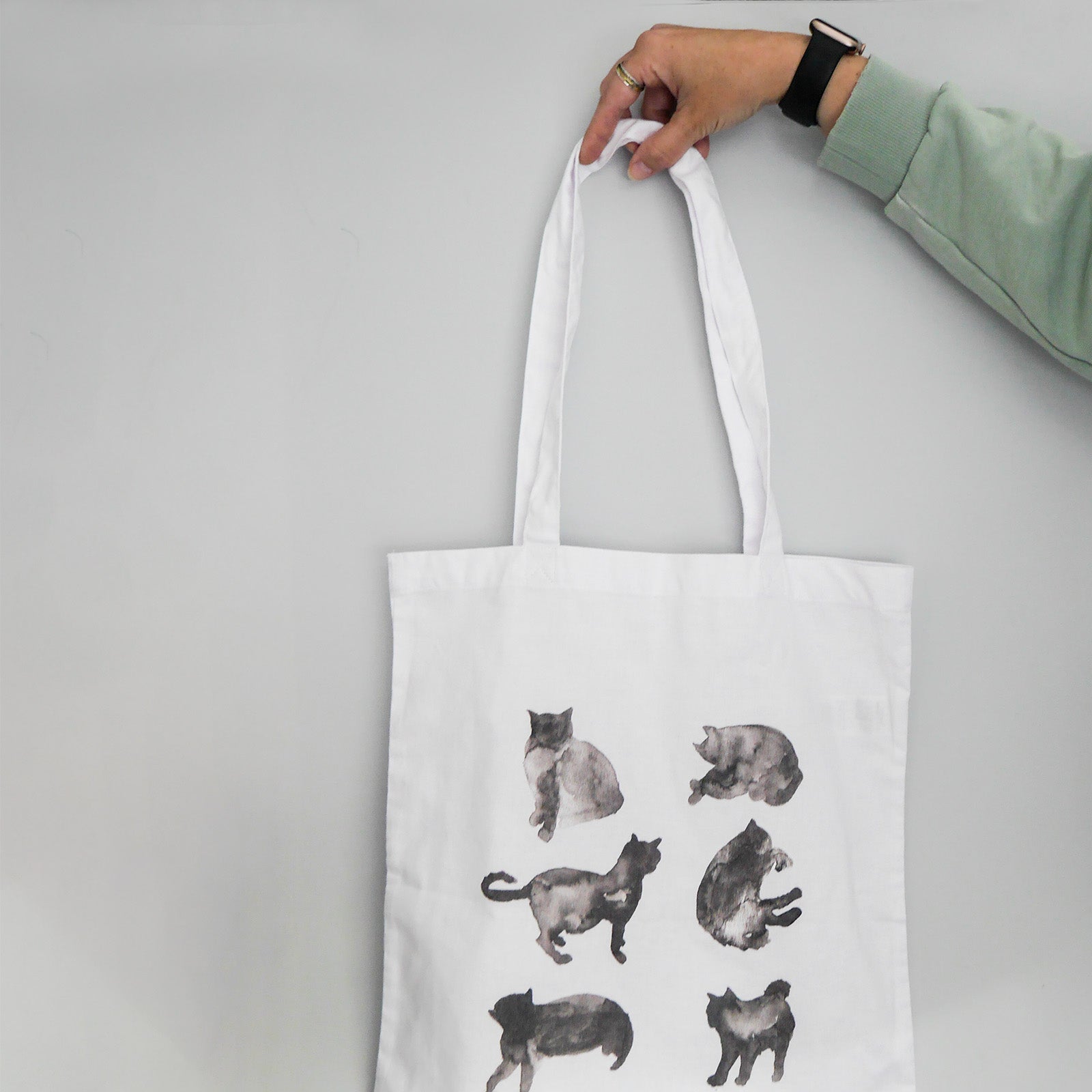 Tote bag with cat illustrations held by a hand on a gray background